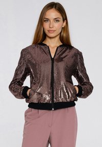Kurtka bomberka w kolorze różowego złota, ozdobiona cekinami, z czarnymi ściągaczami na mankietach i kołnierzu; posiada zamek błyskawiczny z przodu i boczne kieszenie. Gładka, błyszcząca faktura.