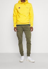 Gul hættetrøje med frontlomme, lilla logo; olivengrøn cargo-bukser med sidelommer; hvide sneakers med minimalistisk design.
