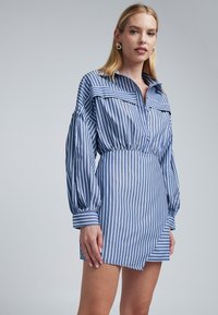 Abito camicia blu a righe con colletto abbottonato, maniche lunghe a palloncino e vita elastica. Presenta un orlo diagonale e una texture morbida.