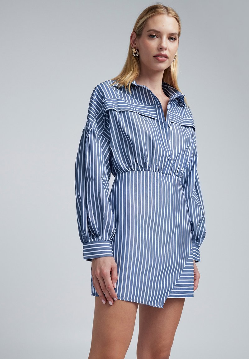 Pruhovaná modrá košilová šaty s knoflíkovým límcem, dlouhými balónovými rukávy a elastickým pasem. Obsahuje diagonální lem a měkkou texturu.