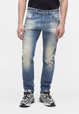 Personne portant un jean bleu délavé et des baskets noires et argentées, debout devant un fond blanc uni.