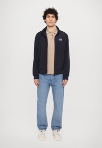 Chaqueta navy con cremallera y alto cuello, llevada sobre una camiseta polo beige, combinada con unos jeans rectos de color azul claro y zapatillas blancas.