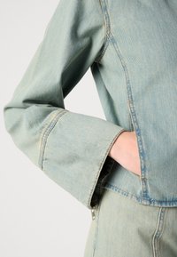 Lys blå denimjakke med en avslappet passform, synlige sømmer og glidelåsdetalj ved mansjetten. Stoffet er mykt med en lett falmet tekstur.