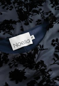 Crna cvjetna čipka na prozirnom tamnoplavom materijalu s glatkim plavim izrezom. Oznaka kaže "Noella" s oznakom veličine "S."