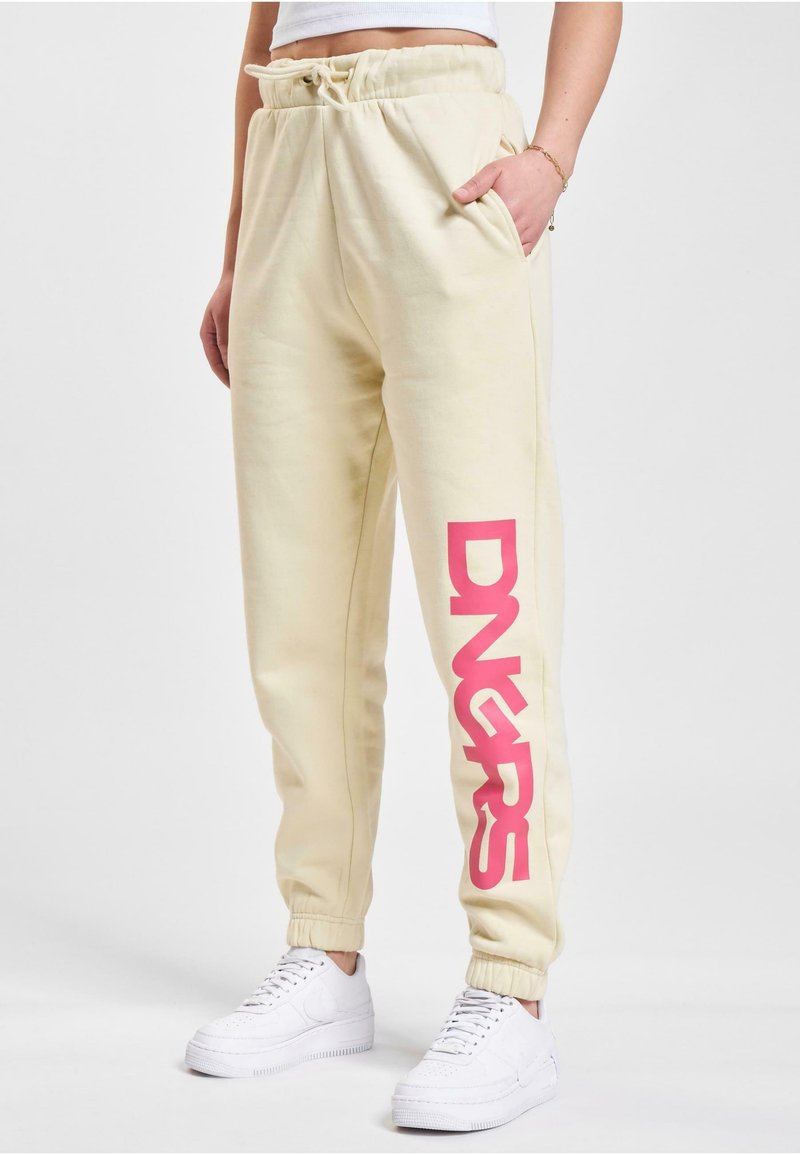 Cremfarbene Sweatpants mit elastischen Bündchen und Bund, die auf dem linken Bein eine große, auffällige pinke "DNGRS" Grafik zeigen.