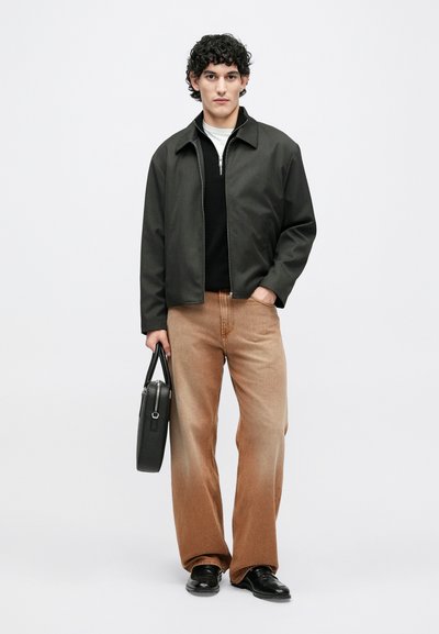 Tmavosivá bunda na zips cez čierny sveter, v kombinácii so svetlohnedými rozšírenými džínsami. Modelka drží čiernu tašku a má na sebe čierne topánky.