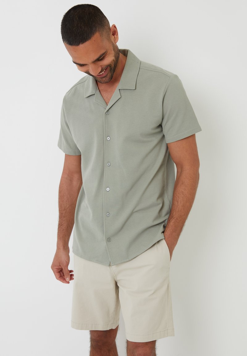 Camisa de mangas cortas de color verde claro con textura suave, que presenta un cierre de botones en la parte delantera y un sutil detalle de costura. Llevada con pantalones cortos beige.