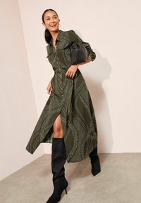 Rochie călcată de culoare verde măslin, cu un model de puncte albe și linii ondulate, cu fante laterale, asortată cu cizme negre din suede până la genunchi și o geantă neagră.