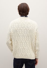 Weißer Strickpullover mit strukturiertem Zopfmuster, rundem Ausschnitt sowie gerippten Bündchen und Saum. Über einem karierten Hemd getragen.