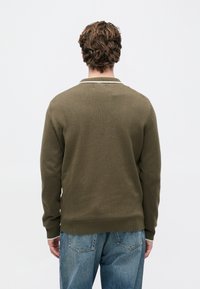 Homem com cabelo castanho encaracolado, vestindo um suéter verde oliva e jeans azuis, de pé virado de costas contra um fundo branco simples.