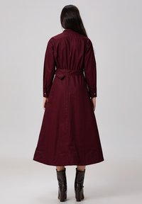 Robe bordeaux à manches longues avec une taille ajustée et des poignets à boutons, dotée d'une ceinture et d'une jupe longue, fabriquée en tissu doux.