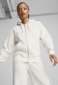 Donna che indossa una felpa con zip in pile color crema e pantaloni abbinati, in piedi al chiuso contro uno sfondo di soffitto e pareti bianche.