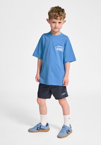 Lichtblauw T-shirt met korte mouwen en een bedrukte logo, gecombineerd met donkerblauwe shorts. De outfit wordt aangevuld met blauwe sneakers en witte sokken.