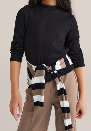 Person iført en sort langærmet top med en stribet sort, hvid og brun sweater bundet omkring taljen, kombineret med brune bukser.