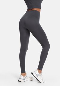 Tmavé šedé legíny s vysokým pásom vyrobené z elastického materiálu, s hladkým povrchom a priliehavým strihom, v kombinácii s bielymi teniskami.