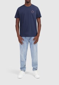 Marineblauwe katoenen T-shirt met korte mouwen, ronde hals en een klein circulair logo op de borst. Gecombineerd met lichtgewassen denim jeans en witte sneakers.