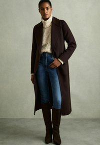 Lungo cappotto marrone, maglione a trecce crema, jeans blu e stivali marroni al ginocchio. Tessuto liscio, design strutturato e silhouette aderente.