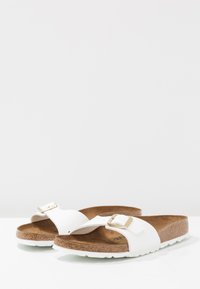 Witte slide-sandalen met een brede band, gouden gespdetails, kurken voetbed en een gestructureerde rubberen zool, met een minimalistisch ontwerp.