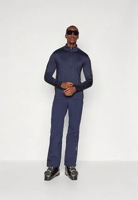 Mees, kes kannab tumesinist suusariietust, musti suuskade saapaid ja suuri musti päikeseprille, seisab heledal ühtlasel taustal.