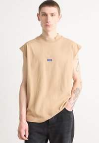 Beige bezrukávové tričko s kulatým výstřihem, opatřené modrým obdélníkovým logem. Tkanina vypadá měkce a má uvolněný střih.