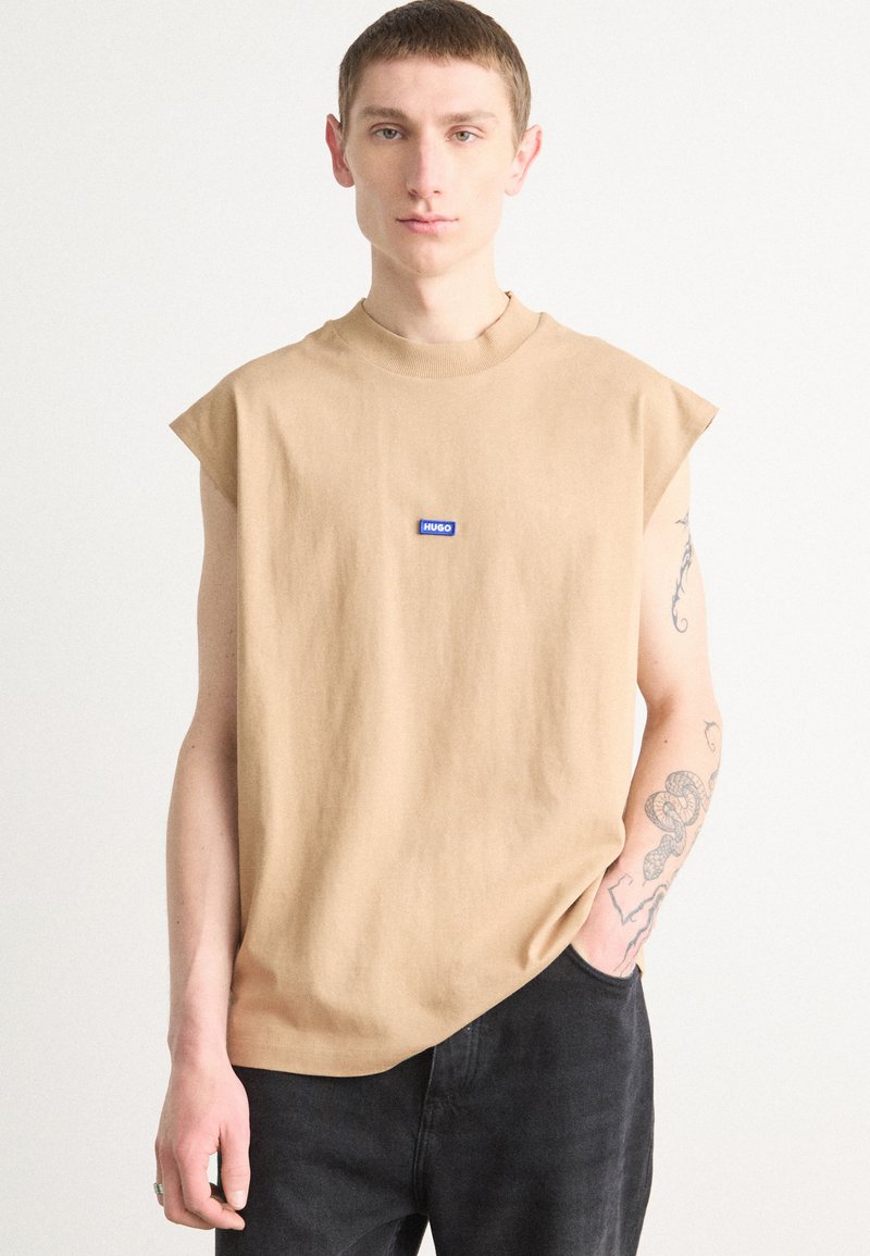 Beige bezrukávové tričko s kulatým výstřihem, opatřené modrým obdélníkovým logem. Tkanina vypadá měkce a má uvolněný střih.