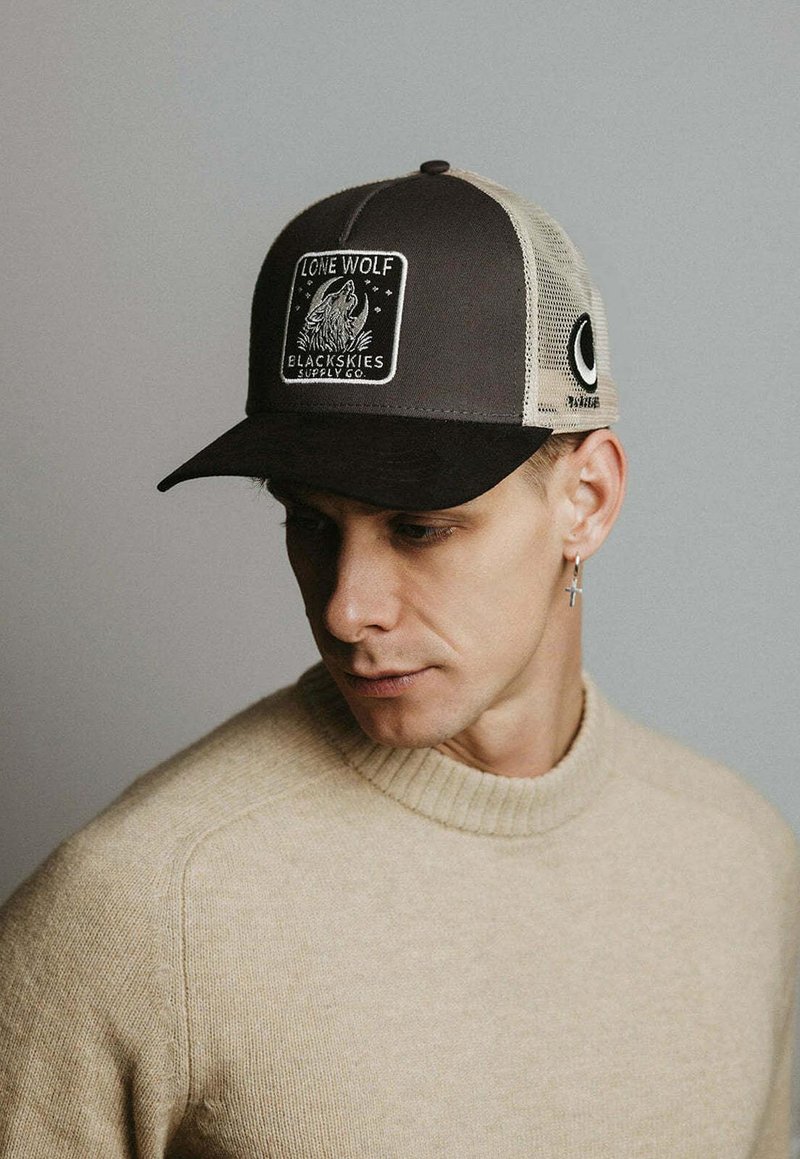 Mladi moški, oblečen v bež pulover in črno-bež kapo z oznako »Lone Wolf Blackskies Supply Co«, gleda navzdol levo.