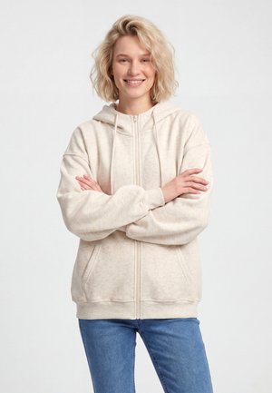 Femme blonde souriante, bras croisés, portant un sweat à capuche beige clair zippé et un jean bleu sur un fond uni.