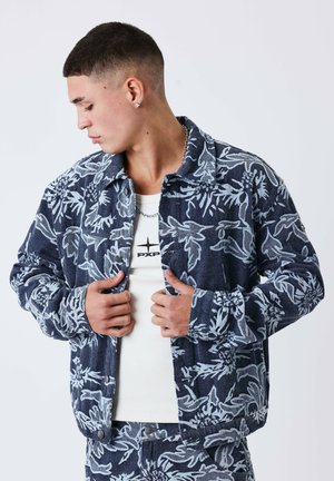 Jeune homme aux cheveux courts portant une veste à motif floral bleu et blanc et un pantalon assorti, tenant la veste ouverte pour révéler un t-shirt blanc.