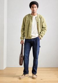 Giacca in pile verde con bottoni a pressione, t-shirt beige, jeans blu con pannelli cuciti e scarpe scamosciate color cammello; borsa a spalla marrone.
