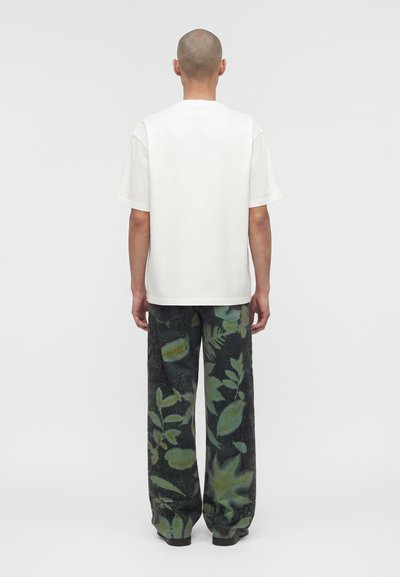 Άτομο με ξυρισμένο κεφάλι που κοιτάζει προς τα πίσω, φοράει απλό λευκό μπλουζάκι με κοντά μανίκια και άνετο σκούρο παντελόνι με πράσινα φυτικά σχέδια.