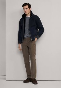 Navy bomber jacket with a high collar, gray textured sweater underneath, brown trousers, and dark brown lace-up shoes.