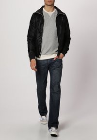 Svart bomberjacka med dragkedja över en ljusgrå tröja, tillsammans med mörkblå jeans och vita sneakers. Har elastiska manschetter och midja.