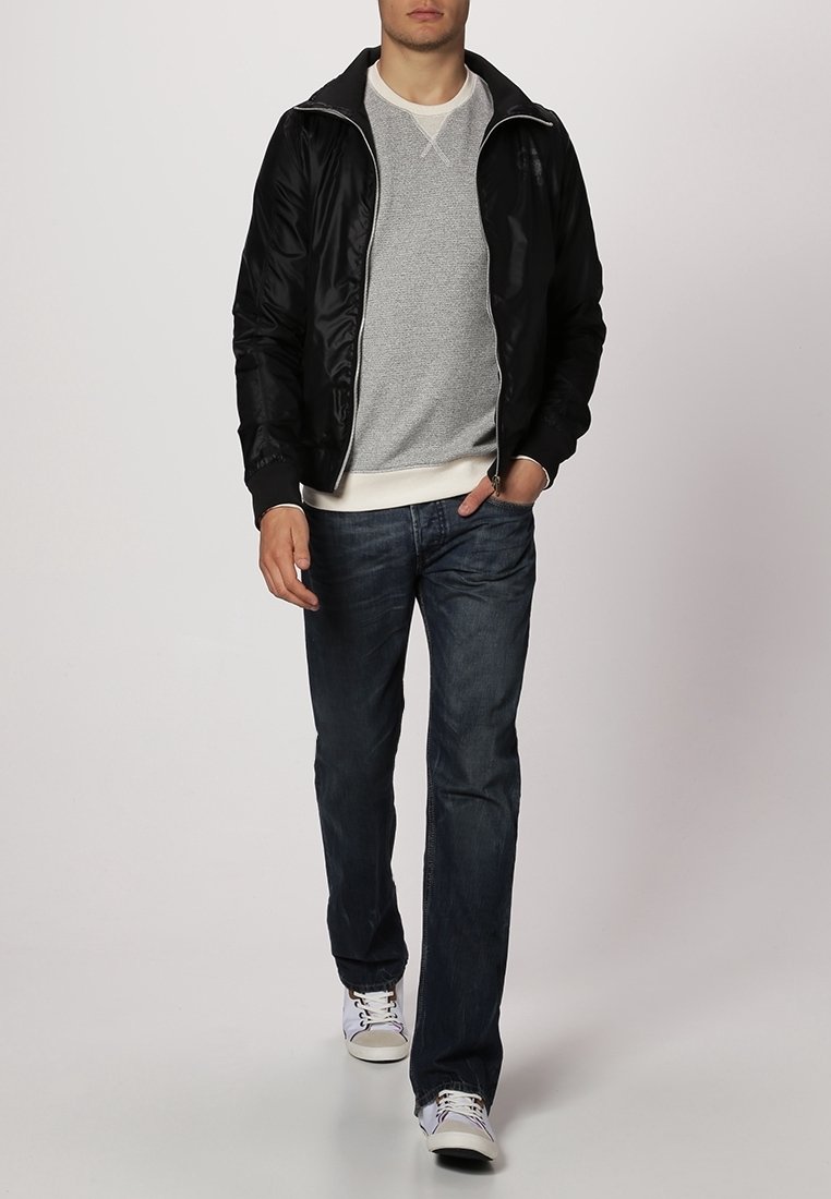 Svart bomberjacka med dragkedja över en ljusgrå tröja, tillsammans med mörkblå jeans och vita sneakers. Har elastiska manschetter och midja.