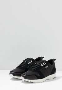 Ett par svarta Hugo låga sneakers med vita sulor och snörning, placerade framåt på en vit yta.