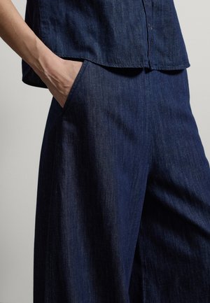 Personne portant un pantalon en jean bleu foncé et une chemise, la main gauche dans la poche du pantalon, sur un fond uni.