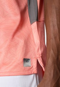 Camiseta deportiva rosa con un patrón geométrico texturizado, detalles grises y una etiqueta de marca en el dobladillo. Tela suave con aberturas laterales.