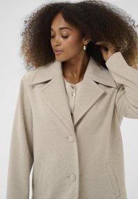 Cappotto beige in misto lana con colletto a rever, due bottoni e tasche laterali; presenta un tessuto testurizzato e una vestibilità comoda.