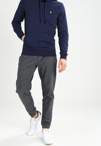 Felpa blu navy con tasca frontale e cordino, abbinata a pantaloni tuta grigi a righe e sneakers bianche con dettagli rossi e blu.