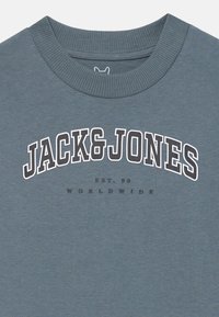 Mėlynas megztinis su apykakle, su raukšlėtu kaklu ir baltu drąsiu tekstu "JACK & JONES" virš mažesnio užrašo "EST. 90 WORLDWIDE" priekyje.