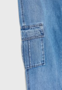 Tela de mezclilla azul claro con un bolsillo lateral tipo cargo. El material tiene una textura suave y presenta costuras amarillas a lo largo de las costuras.