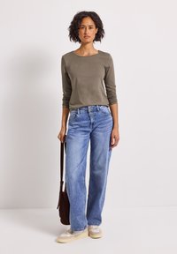 Kvinna med lockigt hår som bär en olivgrön tröja med lång ärm, ljusblå vida jeans, beige sneakers och håller en brun axelväska, stående mot en enfärgad bakgrund.