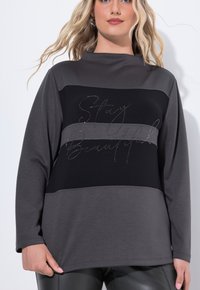 Pull à manches longues avec un design en color-block noir et nuances de gris, avec une inscription métallique "Restez Belle" sur le devant.