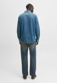 Cămașă din denim de culoare albastru mediu, cu mâneci lungi și manșete cu nasturi, și detaliu de umeri în spate. Asortată cu blugi drepți decolorati și pantofi maro.