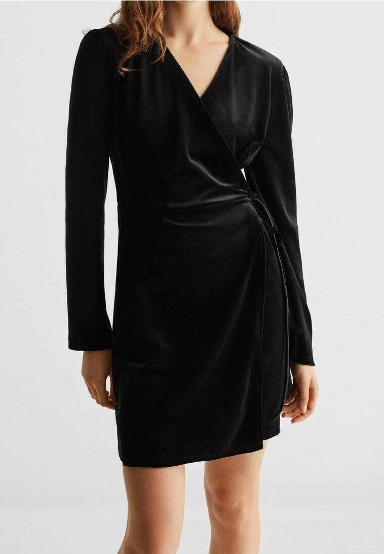 Femme portant une robe portefeuille en velours noir à manches longues, décolleté en V et détail noué à la taille sur un fond clair uni.