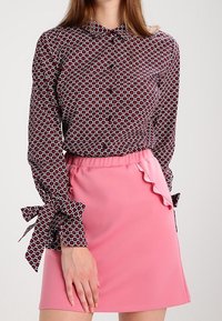 Röd och vit prickig skjorta med knäppning och bandknutna ärmar; parat med en rosa volangkjol i mini-längd.