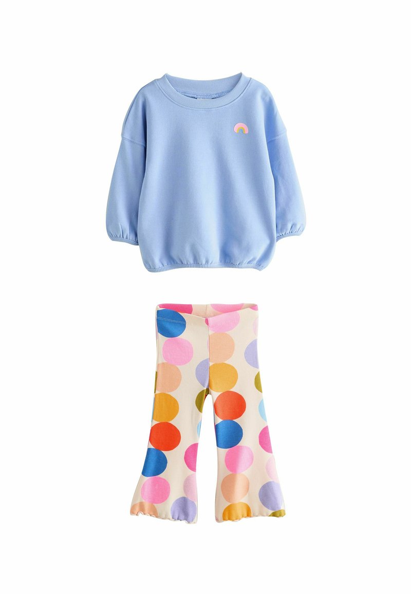 Ljusblå sweatshirt med elastiska ärmslut och ett regnbåge-märke, matchad med krämfärgade leggings med flerfärgade prickar i olika storlekar.