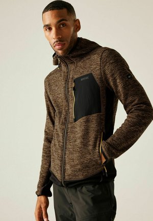 Mężczyzna ubrany w brązową bluzę z kapturem zapinaną na zamek, z czarną kieszenią na piersi i czarne spodnie, stojący z jedną ręką w kieszeni na jednolitym tle.