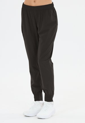 Person iført sorte joggerbukser med elastisk talje og manchetter samt hvide sportssko, stående foran en ensfarvet hvid baggrund.