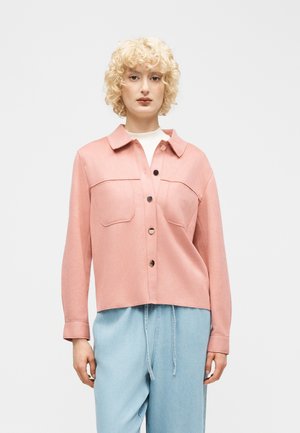 Personne blonde portant une chemise rose clair boutonnée avec des poches poitrine et un pantalon taille haute bleu clair, sur un fond uni.