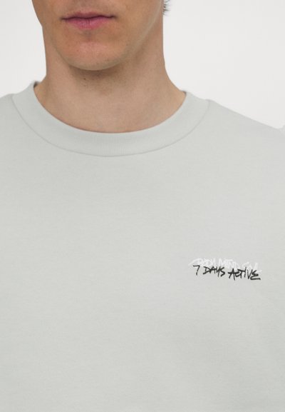 Tricou dintr-un material moale, de culoare gri deschis, cu un decolteu rotund, având un mic text grafic alb-negru pe piept. Textura materialului este netedă.